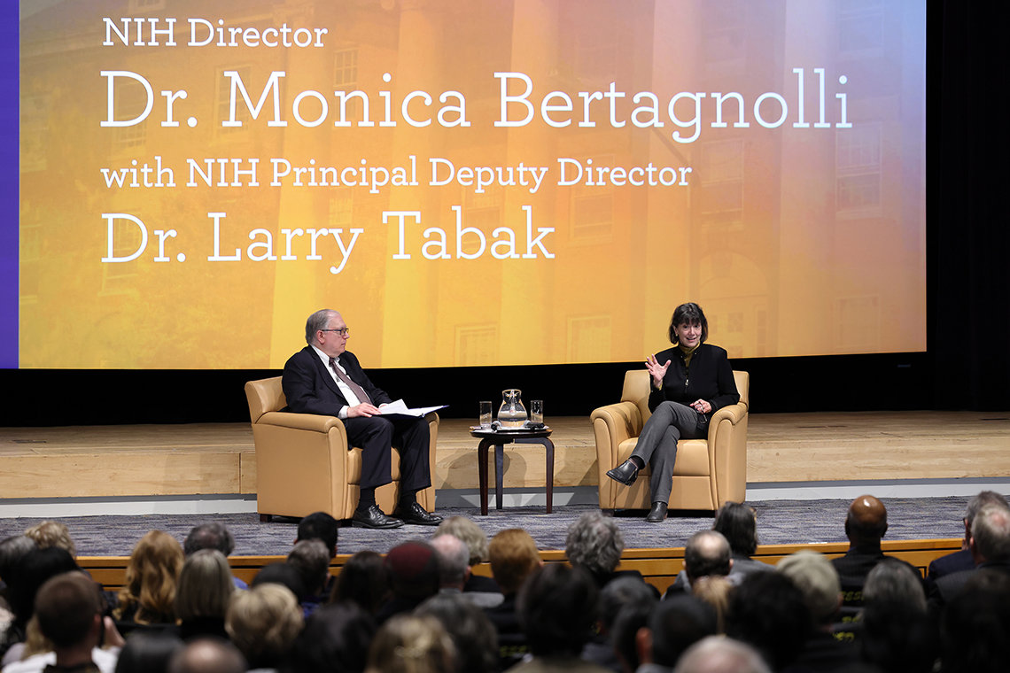 Wide view of Tabak and Bertagnolli sitting in chairs on stage, chatting, with rows of attendees watching