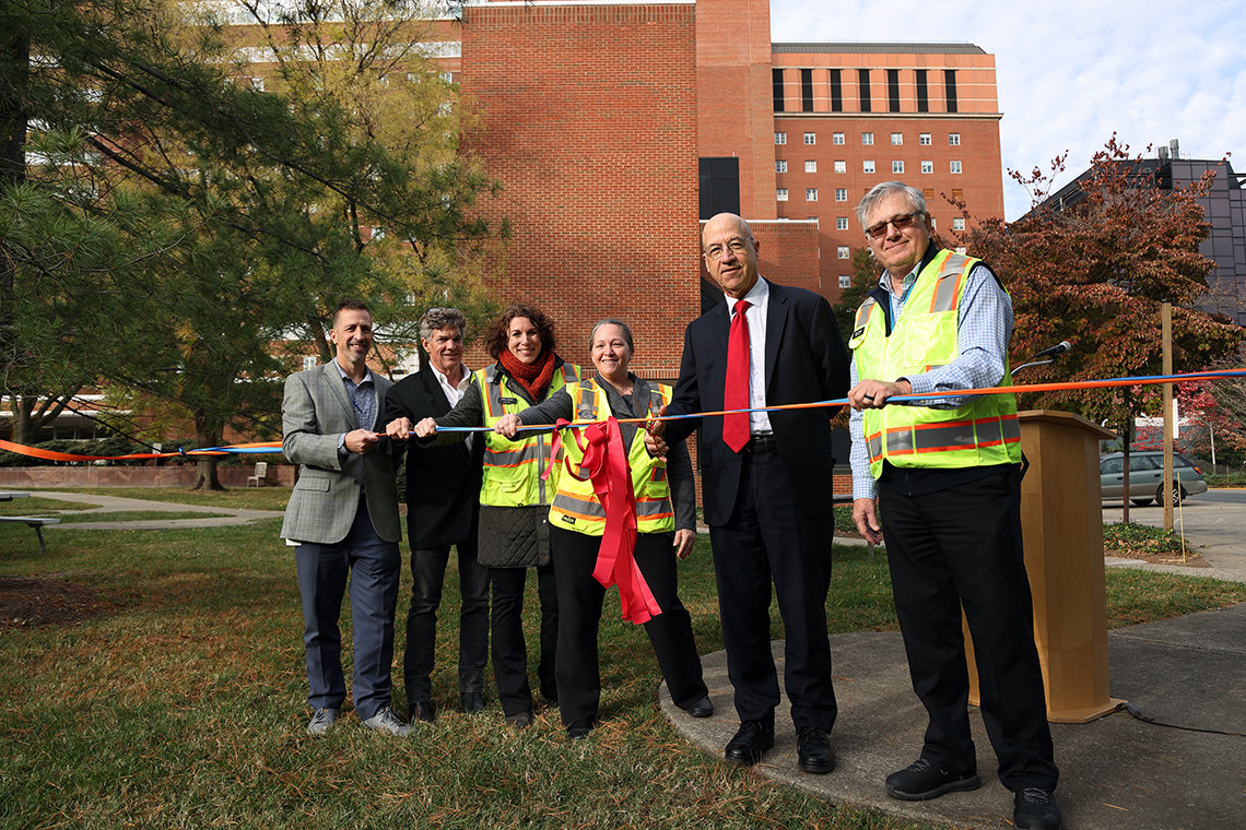 A line of people stand behind a multicolored ribbon. Three people wear suits and three wear fluorescent vests.
