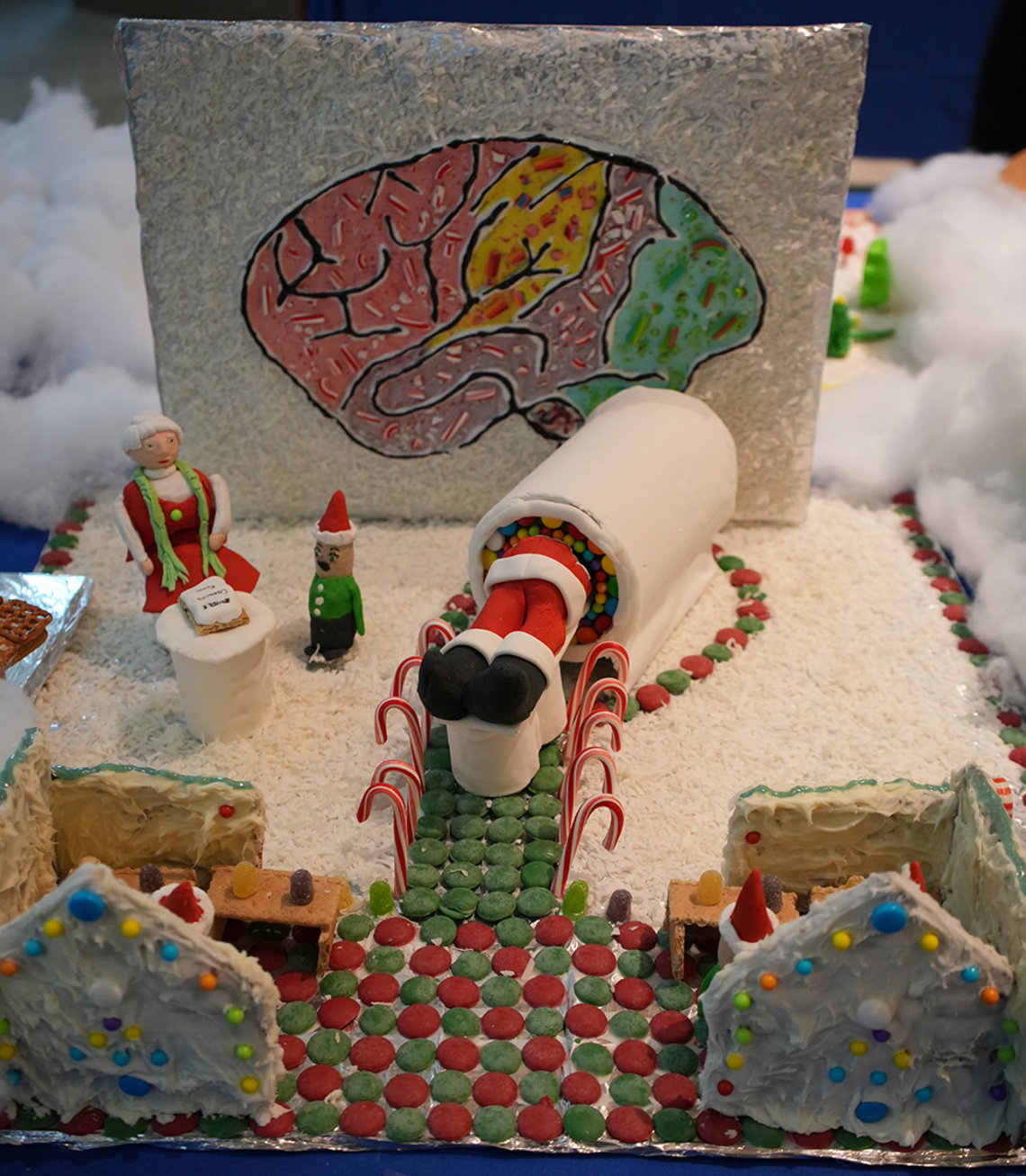 Santas legs stick out from MRI machine, with Mrs. Claus doing intake and a candy brain on display behind