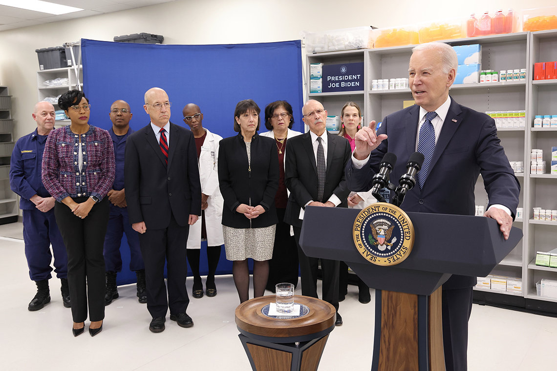 President Biden gesturing from a podium with the presidential seal. Behind him are several men and women standing in a pharmacy room with shelves of medicines visible.