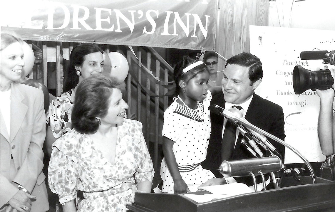 A black and white photo showing a young Broadbent speaking into a microphone as several adults look on.