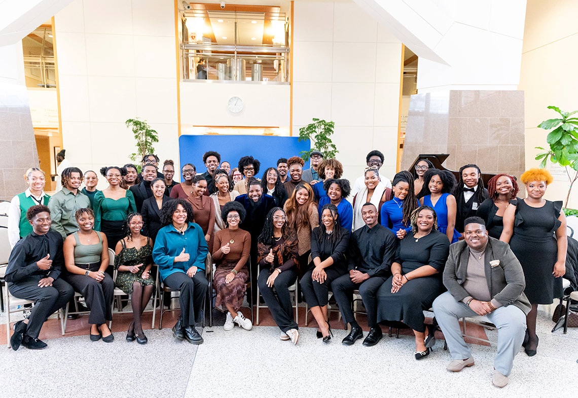 Large group of African American men and women smile for camera with CC atrium behind them.