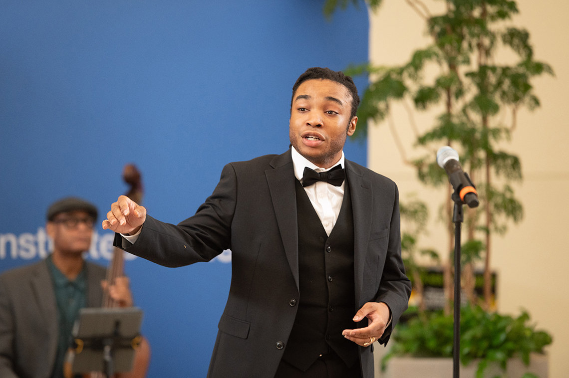 Young Black man in black suit and bowtie gestures as he sings. 