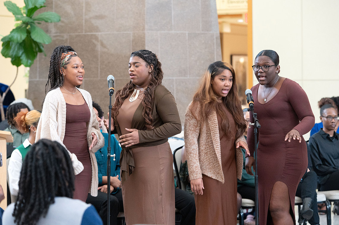 Four young Black women, in pairs, sing into microphone.