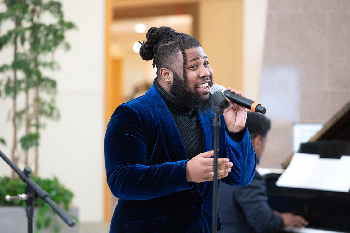 Young Black man wearing a black turtleneck and blue velvet jacket sings into handheld mic.