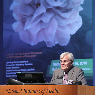Gottesman at NIH podium with 2015 festival slide projected on screen behind him.