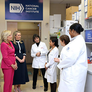 Several women--four in labcoats--chat in a lab environment with NIH NCI logo on wall behind them.