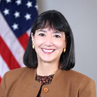 Headshot of Monica Bertagnolli. She is wearing a brown jacket with a patterned red shirt. An American flag takes up the left side of the background.