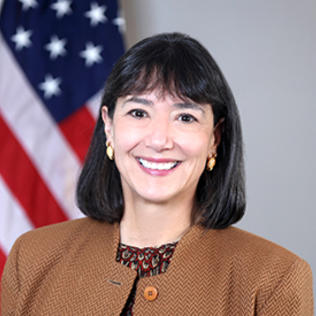 Headshot of Monica Bertagnolli. She is wearing a brown jacket with a patterned red shirt. An American flag takes up the left side of the background.