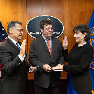 Becerra raise right hand, faces Monica Bertagnolli who has right hand raised. Bertagnolli's husband Alex holds a book, and smiles.