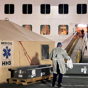 A responder in mask and gown holding a plastic bag walks onto a cruise ship.