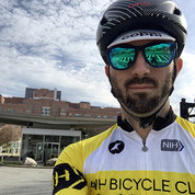 Velez, in bicycle helmet and riding gear, with Clinical Center in background