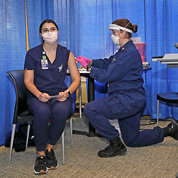A masked who is also wearing a face shield gives another masked nurse a vaccination.