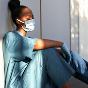 A female nurse in blue scrubs, mask and gloves sits, leaning against a hospital wall, asleep.