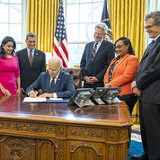 President Biden seated at desk with several people looking over his shoulder