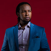 Dr. Ibram Kendi in blue suit, looking off to the side, serious face, against red backdrop.