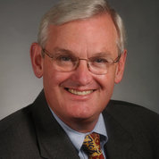 Henderson wears glasses and a black suit, with a blue shirt and gold, black, and red patterned tie. The photo background is gray.