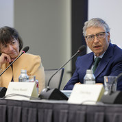Gates and Bertagnolli seated at a long table. Bertagnolli, to the left and slightly out of focus, listens to Gates, who speaks into a microphone while looking to the right and out of the frame.