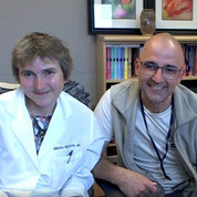 Woman in labcoat beside man wearing glasses, both smiling into camera